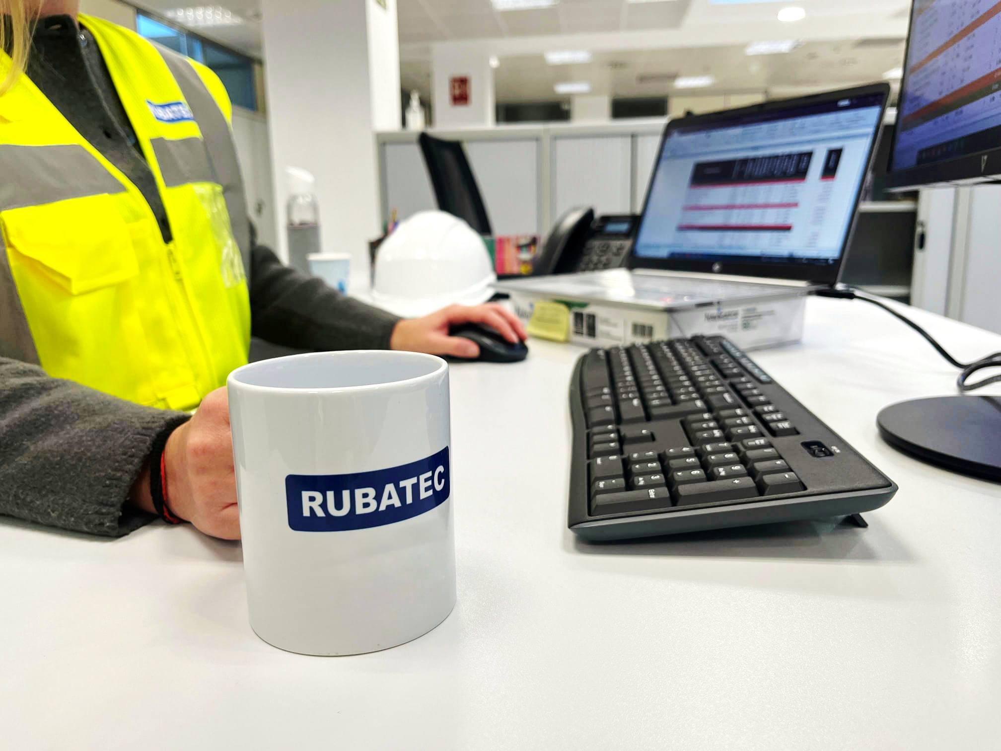 Persona con chaleco reflectante de Rubatec trabajando frente al ordenador en una oficina. En primer plano, una taza con el logotipo de Rubatec.